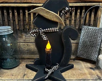 May include: A black wooden cat-shaped Halloween decoration wearing a witch hat and a checkered ribbon. The cat sits on a star-shaped base and holds a battery-operated candle. A vintage jar and grater are in the background.