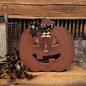 May include: A rustic, wooden pumpkin decoration with a carved face, perfect for Halloween. The pumpkin is painted brown with a black star and a plaid bow. It is displayed on a wooden surface.
