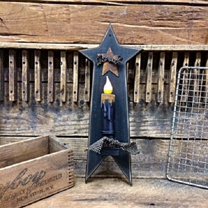 May include: Black wooden star-shaped candle holder with a black and white checkered ribbon and a small star on top. The candle holder has a black candle with a yellow flame.