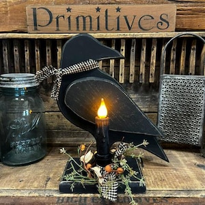 May include: A black wooden crow silhouette figure mounted on a rectangular base, featuring a black taper candle with an orange LED flame. The crow is adorned with a black and white checkered fabric ribbon and a small cluster of artificial greenery and berries.