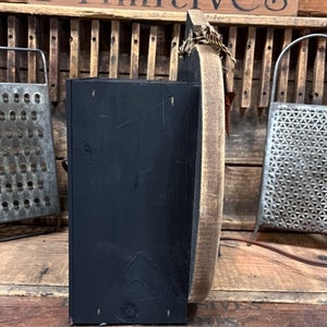 May include: A side view of a black wooden box attached to a circular wooden board with a light brown finish. The box has a matte black surface, and the circular board features a rustic edge. A small piece of twine is tied at the top of the board.