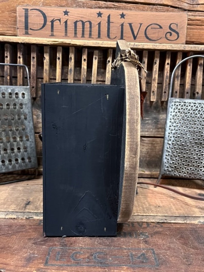 May include: A side view of a black wooden box attached to a circular wooden board with a light brown finish. The box has a matte black surface, and the circular board features a rustic edge. A small piece of twine is tied at the top of the board.