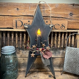 May include: A black wooden star-shaped candle holder with a red and white checkered fabric bow and a sprig of berries. The candle holder is lit with a warm yellow candle.