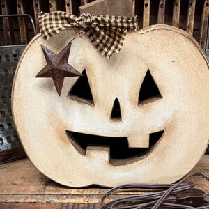 May include: A light-colored wooden jack-o'-lantern decoration with a distressed finish, featuring triangular eyes and a wide, toothy grin. A brown and tan checkered fabric bow sits at the top, accented by a small, rusted metal star hanging on one side.