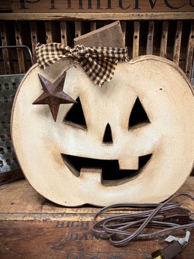 May include: A light-colored wooden jack-o'-lantern decoration with a distressed finish, featuring triangular eyes and a wide, toothy grin. A brown and tan checkered fabric bow sits at the top, accented by a small, rusted metal star hanging on one side.