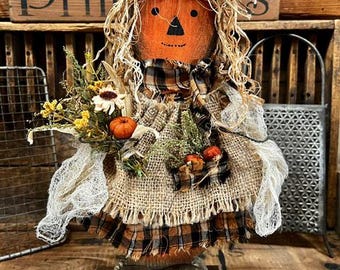 May include: A decorative scarecrow figure with an orange pumpkin head, a black triangular nose, and a stitched mouth. It wears a burlap hat, a plaid collar, and a frayed burlap dress adorned with dried flowers, small pumpkins, and white gauze.