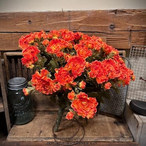 May include: A dense bouquet of vibrant orange and yellow flowers with green stems, arranged in a circular metal spring base. The blossoms feature layered petals in varying shades of bright orange, creating a full, rounded floral display.