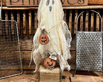 May include: A ghost figurine made of off-white cheesecloth with a hand-drawn face, standing on a square wooden block. It features two small orange pumpkin cutouts with black faces, one hanging from twine and one at the base with a tag reading BOO.