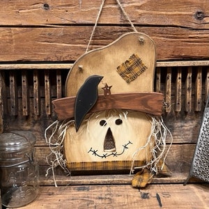 May include: A wooden scarecrow wall hanging featuring a tan hat with a yellow plaid patch, a dark brown brim, and a black crow cutout. The face has black button eyes, a triangular nose, and a stitched mouth, with raffia hair and a plaid fabric trim.
