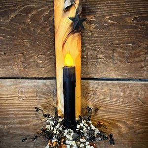 May include: A rustic wooden candle holder with a black candle and a decorative floral arrangement. The candle holder features a black star and a black and white checkered ribbon. The candle has a yellow flame.