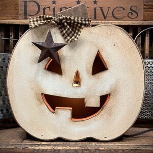 May include: A distressed off-white wooden jack-o'-lantern decoration with a cutout face, a brown and tan checkered fabric bow at the top, and a dark rusted metal star accent. The pumpkin features a warm orange interior glow through the cutouts.
