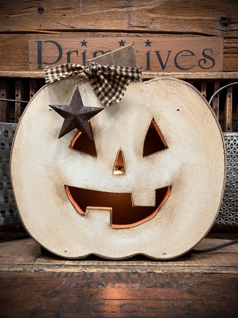May include: A distressed off-white wooden jack-o'-lantern decoration with a cutout face, a brown and tan checkered fabric bow at the top, and a dark rusted metal star accent. The pumpkin features a warm orange interior glow through the cutouts.