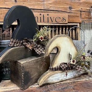 May include: Two wooden witch hats, one black and one cream-colored, are decorated with plaid ribbon and dried flowers. A small wooden box sits between them. The word "Primitives" is visible on a wooden sign in the background.