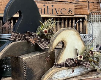 May include: Two wooden witch hats, one black and one cream-colored, are decorated with plaid ribbon and dried flowers. A small wooden box sits between them. The word "Primitives" is visible on a wooden sign in the background.