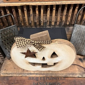 May include: A light-colored wooden pumpkin decoration with a carved face, featuring a brown and tan checkered fabric bow tied around the stem and a small, rusted metal star ornament hanging from the bow, set against a dark wooden background.