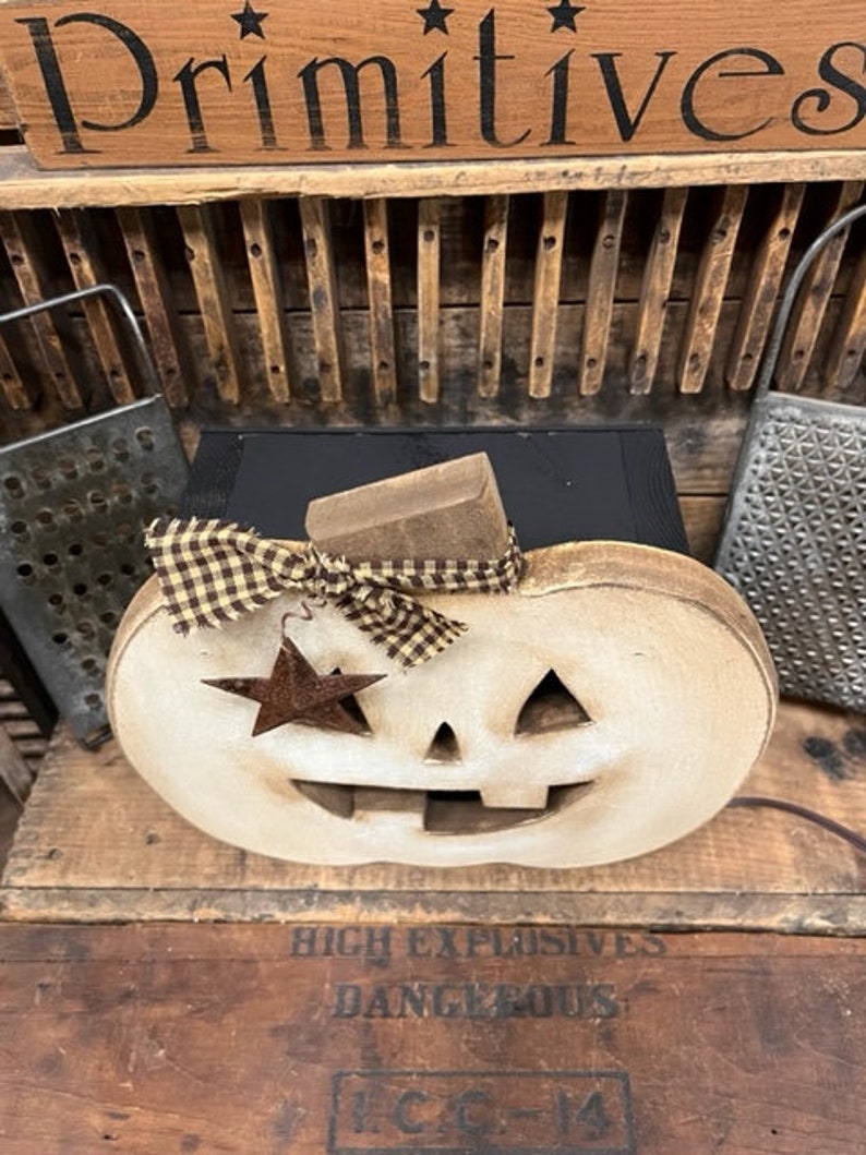 May include: A light-colored wooden pumpkin decoration with a carved face, featuring a brown and tan checkered fabric bow tied around the stem and a small, rusted metal star ornament hanging from the bow, set against a dark wooden background.