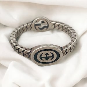 May include: A silver-toned bracelet with a repeating chain design. The bracelet features two oval-shaped centerpieces and a circular centerpiece, each displaying a black logo. The bracelet is displayed on a white fabric.