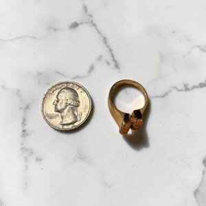 May include: A gold-toned ring with two orange-colored stones, next to a U.S. quarter coin. The ring has a simple band and a decorative setting for the stones. The quarter is silver and shows a profile of George Washington.