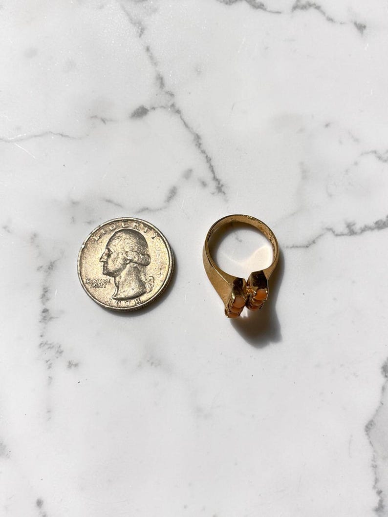 May include: A gold-toned ring with two orange-colored stones, next to a U.S. quarter coin. The ring has a simple band and a decorative setting for the stones. The quarter is silver and shows a profile of George Washington.