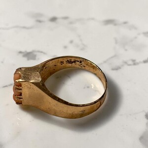 May include: A gold-toned ring with a rectangular face featuring small, coral-colored stones. The ring's band is smooth and rounded, showing signs of wear. The ring is set against a white and gray marble surface.