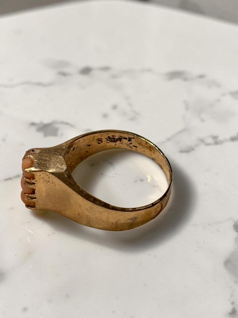May include: A gold-toned ring with a rectangular face featuring small, coral-colored stones. The ring's band is smooth and rounded, showing signs of wear. The ring is set against a white and gray marble surface.