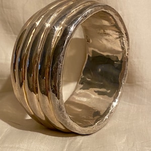 May include: A silver bangle bracelet with a hammered finish and a textured, ridged design. The bracelet is thick and wide, with a rounded shape.