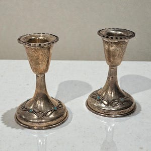 May include: Two silver-toned candle holders with a textured finish. Each holder has a flared top with a decorative edge and a base adorned with turquoise-colored accents. The candle holders are displayed on a white surface.