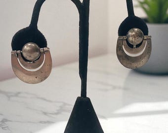 May include: Silver-toned earrings on a black stand. Each earring has a half-moon shape with a sphere. The earrings have a vintage look, with a slightly tarnished finish. The stand is on a white surface.