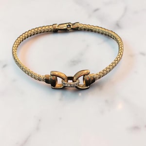 May include: A gold-toned bracelet featuring a woven, mesh-like band and a central metal clasp element. The clasp is shaped like two connected loops with a small, horizontal bar in the center that is accented with several tiny, clear stones.