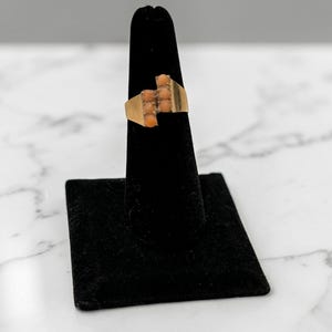 May include: A gold-toned ring with a rectangular design, featuring a series of small, coral-colored stones. The ring is displayed on a black stand against a white and gray marble background. The ring's design is geometric and symmetrical.