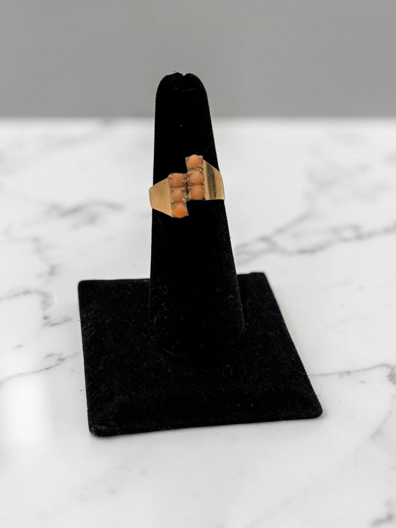 May include: A gold-toned ring with a rectangular design, featuring a series of small, coral-colored stones. The ring is displayed on a black stand against a white and gray marble background. The ring's design is geometric and symmetrical.