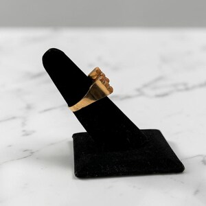 May include: A gold-colored ring with a textured design is displayed on a black velvet ring holder. The ring has a rectangular band and a cluster of textured elements on the face. The background is a marble surface.