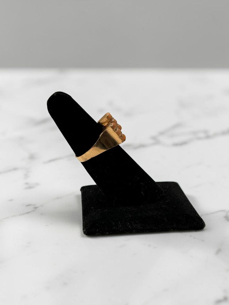 May include: A gold-colored ring with a textured design is displayed on a black velvet ring holder. The ring has a rectangular band and a cluster of textured elements on the face. The background is a marble surface.