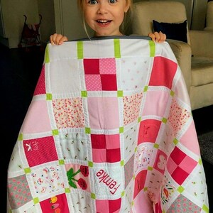 May include: A white and pink patchwork quilt with green trim. The quilt is made up of squares of different fabrics, including floral prints, polka dots, and solid colors. The quilt is personalized with the name "Emma" embroidered in the center.
