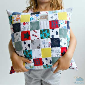 Könnte beinhalten: Ein Patchwork-Kissen mit verschiedenen bunten Stoffquadraten mit unterschiedlichen Mustern, darunter Anker, Sterne, Schildkröten und Wale. Das Kissen wird von einem Kind gehalten.