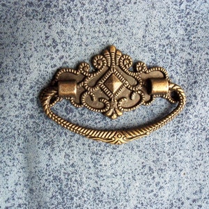 May include: A decorative, ornate, brass-colored drawer pull with a diamond-shaped center and a rope-like design around the edge.