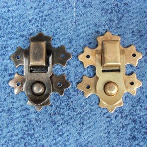May include: Two decorative metal clasps. One is a dark bronze color, and the other is gold. Each clasp has a rectangular latch and a decorative, star-shaped design with a circular center. The clasps are set against a blue background.