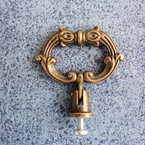 May include: An antique bronze-toned drawer pull with an oval handle and ornate detailing. The handle is attached to a mounting bracket and a screw. The hardware is designed for furniture restoration or decorative projects.