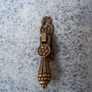 May include: A gold-toned metal drawer pull with an ornate design. The pull has a round knob with a floral pattern and a long, tapered handle.