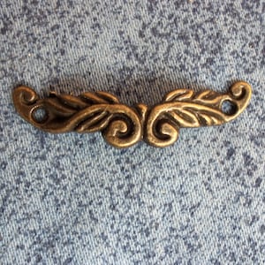 Puede incluir: Una moldura decorativa de metal dorado con un diseño de hojas en espiral. La moldura tiene dos agujeros en cada extremo para sujetarla a un proyecto.