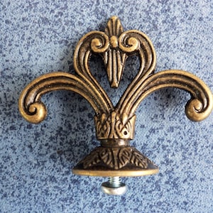 May include: A gold-toned metal decorative finial with a fleur-de-lis design. The finial has a threaded base for attaching to furniture or other objects.