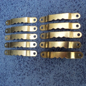 Könnte beinhalten: Zehn goldfarbene Metallbilderhaken mit Sägezahnkanten und zwei Löchern an jedem Haken.