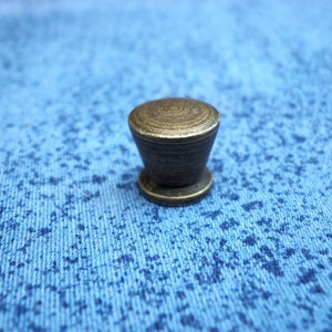 May include: A small, round, brass cabinet knob with a textured surface. The knob is a dark gold color.