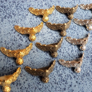 Może przedstawiać: Kolekcja ozdobnych metalowych narożników w odcieniach złota, brązu i srebra. Narożniki mają ozdobny, zakrzywiony kształt i są ułożone na niebieskiej powierzchni materiału.