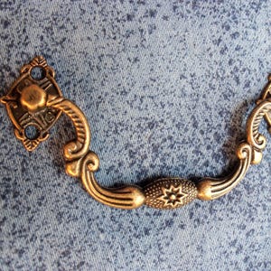 May include: A bronze-toned metal drawer pull featuring a curved, ornate handle with a central textured oval element displaying a star pattern. The handle is attached to two diamond-shaped mounting plates, each with two circular screw holes.