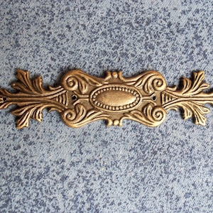 Puede incluir: Adorno decorativo de metal color bronce para muebles. La pieza presenta un diseño ovalado central con un borde de cuentas, flanqueado por volutas simétricas y motivos de hojas. Tiene dos pequeños orificios para el montaje.