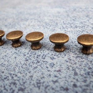 May include: Five antique-style brass knobs with a rounded top and a short stem. The knobs are arranged in a row on a blue textured surface.