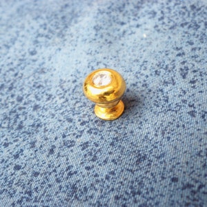 Puede incluir: Un pomo de armario dorado con un strass transparente en el centro.