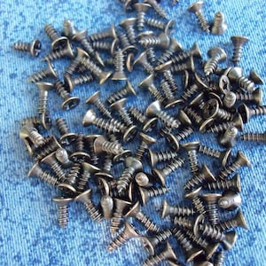 May include: A collection of small, dark metal screws with flat heads and threaded shafts. The screws are scattered on a blue, textured surface, likely fabric. The screws appear to be new and unused, suitable for various construction or repair projects.