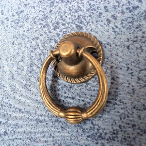 May include: Antique brass-toned drawer pull with a ring handle. The pull has a decorative, ridged design around the circular base and on the handle. The hardware is designed for furniture and has a vintage aesthetic.